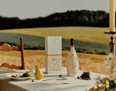 A picturesque rural setting features a table adorned with a white cloth, holding a wrapped bottle, a matching box, and a golden pear. Nearby, blueberries and grapes rest alongside a small plate of nuts. A lit candle stands elegantly on the right. Amidst rolling fields and soft sunlight, the scene exudes rustic charm and tranquility.