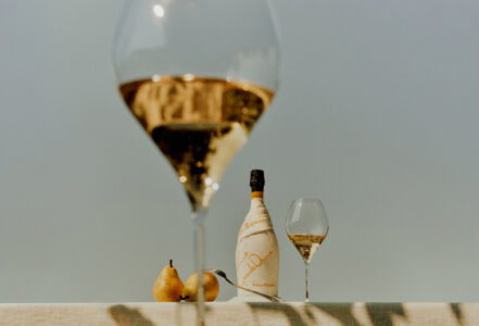 A large wine glass in the foreground holds a sparkling beverage, creating a blurred, artistic effect. In the background, a bottle wrapped in cloth sits alongside a smaller, partially filled wine glass. Two ripe pears rest on a light-colored tablecloth, casting gentle shadows. The scene exudes elegance and simplicity, bathed in soft, natural light.