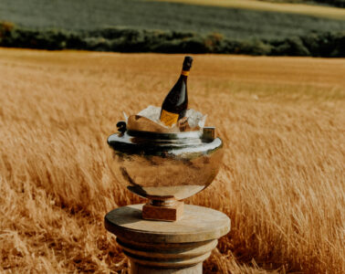 A golden-hued field stretches under a vast sky, with a silver wine cooler prominently placed in the foreground, resting on a pedestal. Inside the cooler, a bottle of champagne sits nestled among ice cubes. The pastoral landscape, with its lush, expansive fields and distant trees, creates a serene, picturesque backdrop for the elegant setting.