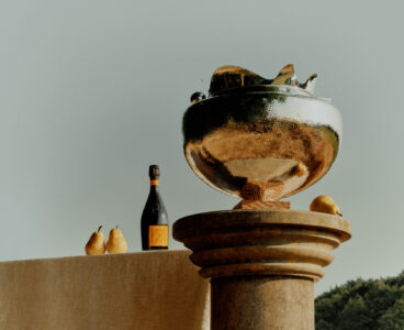 A polished bronze sculpture sits atop a classical stone pillar, reflecting light and surrounding scenery. A beige cloth-covered table holds two ripe pears and a bottle of sparkling wine, positioned in front of a distant, verdant forest. The composition blends elements of nature and still life against a soft, muted sky, creating a serene, elegant atmosphere.