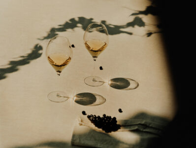 Two wine glasses filled with a golden liquid are elegantly placed on a light-colored surface, casting distinct shadows. A few dark berries are scattered nearby, and more berries rest on a folded napkin in the foreground. Soft shadows from foliage create an artistic play of light and form, adding a touch of nature's grace to the scene.