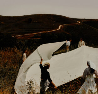 Several individuals are collaboratively spreading a large white sheet outdoors. They are standing in tall grass, and the setting features rolling hills with winding paths in the background. The atmosphere is serene, and the scene suggests teamwork and harmony with nature. The attire of the individuals varies, adding a touch of diversity to the gathering.