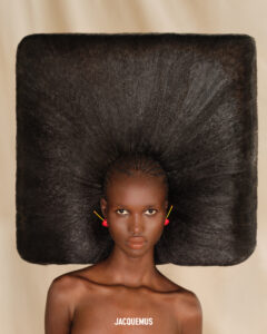 A person displays a striking and unique hairstyle, featuring a massive square-shaped afro that extends horizontally, surrounding the head. The hair radiates from a central point, with intricate braiding visible at the top. The person wears vibrant yellow and red spherical earrings. The background is a soft beige, emphasizing the boldness and creativity of the hairstyle.