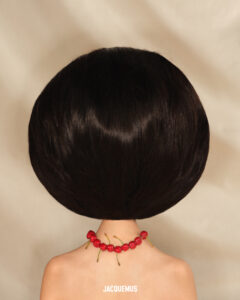 A striking composition features a person with voluminous, sleek dark hair seen from behind, creating a dramatic silhouette. Their shoulders are bare, adorned with a necklace crafted from vibrant red cherries, adding a playful and contrasting touch of color. The neutral background enhances the chic and artistic presentation, focusing attention on the unusual and creative styling elements.