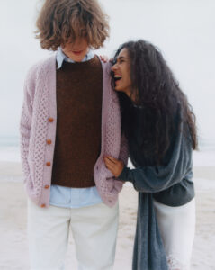 Two people are on a beach, sharing a light-hearted moment. One wears a brown sweater under a light pink cardigan, and the other, in a dark, long-sleeved top, leans in laughing. Both are dressed in light-colored bottoms. Their relaxed posture and the serene background suggest a casual, joyful atmosphere amidst the natural backdrop of sand and sea.