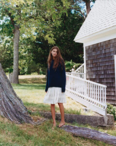 A person with long brown hair stands barefoot on a grassy area near a large tree trunk. They are wearing a dark blue sweater and a white skirt. In the background, there is a white-railed wooden structure with shingles, surrounded by lush green trees and a bright, sunny sky. The scene conveys a serene, rural setting.