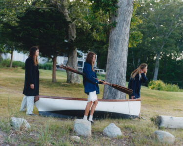 Three individuals are positioned in a grassy area near trees, each wearing different outfits. They appear to be carrying a small, upside-down boat with paddles inside. Stones and shrubs are scattered on the ground, and a large tree stands prominently nearby. The setting suggests a peaceful, outdoor environment, likely near a shore or wooded area.