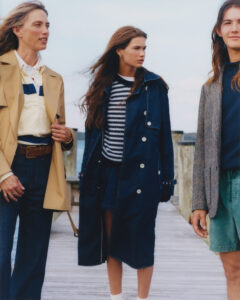 Three individuals stand on a wooden dock under a clear sky, exuding a relaxed yet stylish vibe. On the left, a person wears a beige trench coat and blue jeans, while the center individual sports a navy coat over a striped sweater. The third person on the right combines a tweed jacket with green shorts, creating an eclectic ensemble.