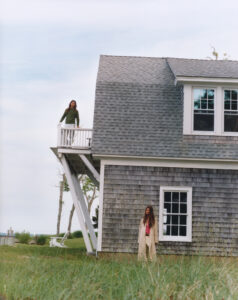 A quaint house with weathered shingles stands in a grassy area. A person in a green top leans on the white railing of a balcony, while another in a beige coat stands below near a window. The scene conveys a serene, coastal vibe with distant trees and a clear sky enhancing the tranquil atmosphere.