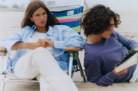 Two individuals relax on a sandy beach. A person wearing a light blue shirt and white pants sits on a striped chair, looking thoughtful. Beside them, another in a purple sweater reclines on the sand, engrossed in a book. The atmosphere appears calm and serene, with the ocean and distant horizon barely visible through mild haze.
