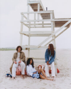 Three individuals are casually positioned under a tall, white lifeguard stand on a sandy beach. The muted, cloudy sky adds a soft backdrop. One person sits barefoot on a buoy, dressed in light clothing, while another leans against the structure. The scene captures a relaxed, summery atmosphere with the ocean and distant greenery visible in the background.