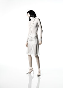 A person stands gracefully against a stark white background, wearing a form-fitting, long-sleeved top and a knee-length skirt, both in soft, light hues. Their sleek bob hairstyle complements the minimalist appeal. The ensemble is accentuated with long, layered necklaces and elegant high-heeled shoes, creating a sophisticated and modern aesthetic with an air of timeless fashion.