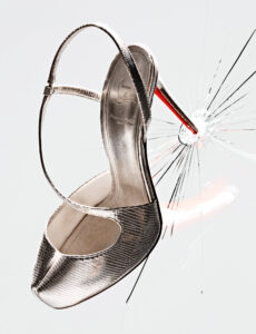 A striking metallic high heel with an elegant strap design is positioned dramatically with its heel penetrating a surface, causing a vivid, radiating crack pattern. The shoe's underside reveals a bold red sole, contrasting with the surrounding light tones. The scene conveys a sense of power and elegance, emphasizing both the shoe's sophistication and its impact.