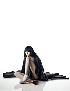 A person with a short black hairstyle sits on a glossy white surface, wrapped in a flowing black garment. One leg is extended, wearing a glittery high heel, while the other shoe lies on its side in front. The expression is pensive, and the dramatic contrast emphasizes the sleek, sophisticated elegance and minimalist aesthetic.