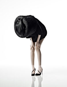 A person in a striking pose bends forward, hands resting on their knees, with their head obscured by a large, woven black hat. They wear a sleek black outfit and shiny high heels, standing against a minimalist white background. The image emphasizes fashion and form, combining elegance with avant-garde elements to create a bold visual statement.