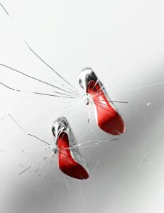 Two high-heeled shoes with silver exteriors and striking red soles appear to pierce through a glass surface, creating spiderweb-like cracks. The background is a soft gradient of white to gray, emphasizing the dramatic contrast of the shoes' colors. The overall effect is both elegant and dynamic, highlighting the bold design of the sleek footwear.