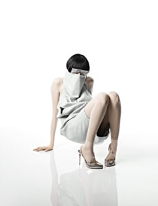 A person with sleek black hair and a futuristic vibe sits on a glossy surface. They wear a light, draped outfit that partially covers their face. Their legs are adorned with light-colored stockings and metallic high heels. The scene exudes a minimalist, avant-garde aesthetic, with a focus on fashion and mystery against a bright, clean backdrop.