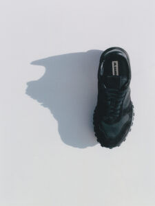 A single black sneaker with a rugged, textured sole is positioned on a smooth, light background. The shoe's laces are tightly tied, and the material appears soft, possibly suede or leather. A stark shadow stretches out to the left, contrasting with the clean, minimalistic space. The overall composition is balanced and stylish, emphasizing simplicity and elegance.