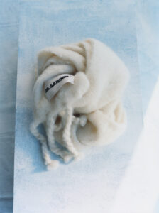 A soft, woolen item sits elegantly on a textured surface, exuding warmth and comfort. The creamy, off-white hue accentuates its cozy appearance, with loosely spun fibers cascading naturally. The gentle play of light and shadow enhances the tactile quality, creating an inviting and serene atmosphere. It suggests a sense of luxury and understated sophistication.