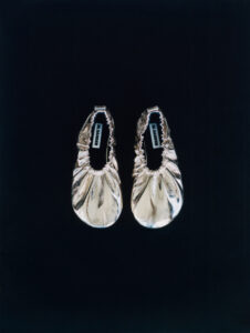 A pair of gold, metallic ballet flats are set against a dark background. The crinkled, glossy material reflects light, creating a luxurious appearance. The shoes have a ruched detailing along the edge and a soft, rounded toe, emphasizing elegance and sophistication. Positioned symmetrically, they convey a sense of simplicity and understated glamour suitable for various occasions.