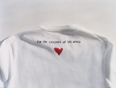 A white T-shirt is displayed from the back, showcasing a simple design. Below the neckline, there is a vivid red heart drawn with a casual style. The material of the shirt appears soft, and the simplistic design conveys a message of warmth and care. Light shadows suggest a natural, relaxed setting, emphasizing the garment’s simplicity.