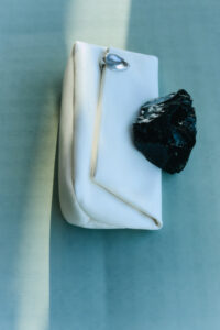 A smooth, white rectangular object rests on a soft, light blue surface. It features a sleek, metal clasp on one corner. A glossy black rock with uneven surfaces lays partially on top of it, contrasting with the polished, minimalist background. A beam of light casts a subtle shadow across the scene, enhancing its serene atmosphere.
