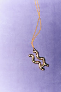 A curvy metallic pendant hangs from a thin, golden chain against a soft purple background. The pendant's reflective surface catches the light, creating glimmers that highlight its unique, wave-like design. The simple yet elegant form contrasts with the understated background, emphasizing the pendant's artistic and organic shape, accentuated by the delicate drape of the chain.