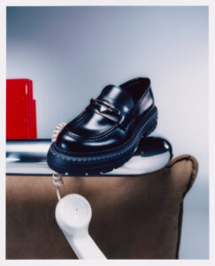 A polished black loafer with a chunky sole rests on a reflective metallic surface. Below, a vintage white telephone handset with a spiraled cord hangs over the edge. In the background, a soft brown cushion contrasts with a striking red block, creating a minimalist yet intriguing composition, blending elements of fashion and retro technology in a modern setting.