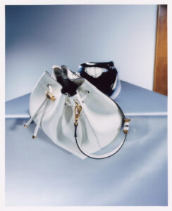 A stylish white handbag with gold accents and sleek black straps is artfully placed on a reflective surface. Its open design reveals a soft, flexible structure, elegantly contrasting with a glossy, abstractly patterned bowl behind it. The setting is minimalistic, featuring soft, muted colors that emphasize the sophisticated and modern aesthetic of the objects.