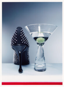 A black stiletto heel adorned with sparkling studs stands beside an elegant martini glass, which holds a single green olive. The sleek design of the shoe contrasts with the smooth curvature of the glass, both displayed on a simple surface with a gradient background, creating a stylish and sophisticated modern composition.