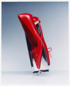 A pair of vibrant red high-heeled shoes, crafted from shiny material, are stylishly displayed against a smooth, muted blue background. The shoes are positioned upright, with delicate red laces adding a playful touch. They rest upon a gleaming, metallic surface that contrasts with their bold color, creating an engaging visual interplay between the different textures and hues.
