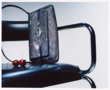 A shimmering black handbag with a sparkling texture is elegantly placed on a sleek, black chair with chrome arms. Beside the bag, two ripe cherries add a pop of color, creating a stylish and sophisticated contrast against the modern design of the chair. The scene exudes luxury and minimalism, blending fashion with simplicity.