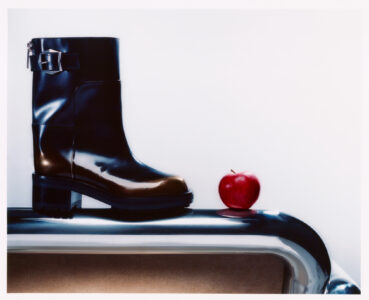 A polished black boot with a decorative buckle rests on a shiny metallic surface, showcasing its sleek design. Beside it, a vibrant red apple adds a pop of color, contrasting with the glossy reflections. The scene combines elements of fashion and nature, emphasizing simplicity and elegance through the juxtaposition of the boot's structured form and the apple's organic shape.