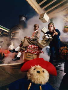 A whimsical scene features a person in plaid attire with colorful braided hair sitting beside a steam engine amidst billowing smoke. Nearby, another person in vintage clothing appears to juggle tumbling boxes. A plush bear in a red hat and blue coat stands prominently in the foreground. The atmosphere is lively and reminiscent of a bustling train station.