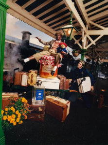 A person dressed in vintage plaid clothing sits atop a stack of luggage at a train station, holding a small stuffed animal. Steam is visible behind, emanating from a classic black locomotive. An attendant in a blue uniform stands nearby, carrying a large box. The scene is vibrant, with bright marigolds adding color to the setting.