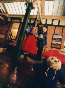 A scene in a vintage-style train station features a woman in a red tulle skirt holding a gift bag, and a man wearing a blue uniform with a suitcase. A small teddy bear in a blue coat and red hat appears at the forefront. The setting includes brick walls, a prominent clock, and old-fashioned luggage, creating a nostalgic atmosphere.