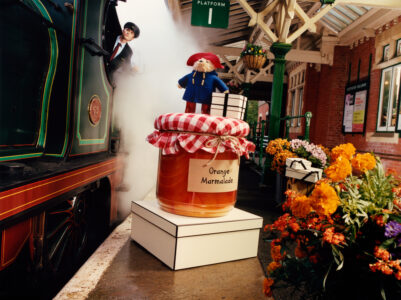 A steam locomotive with a driver emerges onto a railway platform, surrounded by vivid floral decorations. A plush figure in a blue coat and red hat stands beside an oversized jar of orange marmalade on white boxes. The platform is bustling with vibrant colors and architectural details, evoking a nostalgic, whimsical scene.