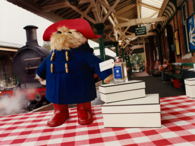 A furry toy wearing a blue coat and red hat stands on a checkered surface, surrounded by stacked white boxes. A glass bottle is placed atop one of the boxes. In the background, a vintage train emits steam at an old-fashioned, covered railway platform featuring green benches and brick walls, evoking a nostalgic and whimsical atmosphere.