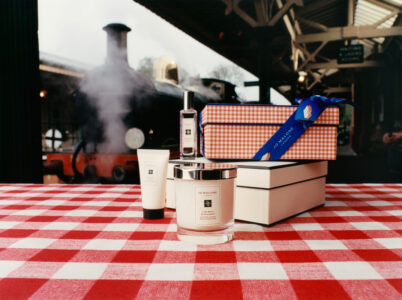 A vintage train emits steam at a station, blending heritage aesthetics with luxury items displayed on a red and white checkered tablecloth. A prominent candle, a tube, and a small bottle share the scene with stacked gift boxes tied with a blue ribbon. The juxtaposition evokes a sense of elegance and timelessness. The background features ironwork and distant trees.