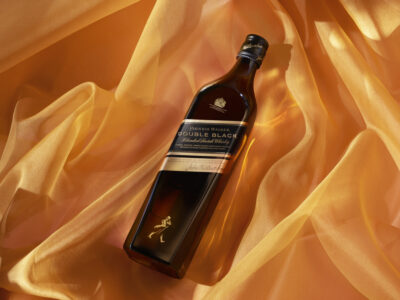 A dark glass bottle lies elegantly on a softly draped, golden-yellow fabric. The bottle's sleek design is accented by golden and black labels, exuding a sense of sophistication and luxury. The warm tones of the fabric create a rich, inviting backdrop, enhancing the bottle's deep hues and giving the scene an indulgent, refined atmosphere.