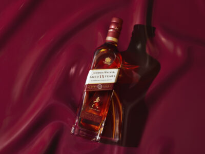 A sleek bottle sits elegantly against a luxurious, deep red, flowing background. The bottle contains a rich, amber liquid and features a golden emblem near the top. Light plays across the bottle's surface, creating highlights and soft reflections that enhance its opulent appeal. Shadows cast an artistic touch, adding depth to the overall composition.