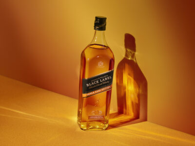 A bottle with a dark cap and label stands elegantly on a smooth, textured surface. The surrounding ambiance is a warm gradient of yellow and orange hues. Sunlight casts a distinct, elongated shadow of the bottle on the background, enhancing its sleek contours and the richness of the liquid it contains.