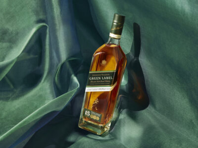 A bottle with a dark green label stands at an angle, casting a shadow on the textured green fabric beneath. The clear liquid inside is rich amber, catching light. The fabric drapes elegantly, creating a soft backdrop. Golden highlights on the bottle add a touch of luxury, enhancing its premium appearance amidst the lush, flowing material.