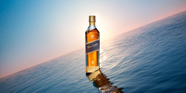 A bottle stands majestically on a calm ocean surface, reflecting in the water. The scene is set against a serene backdrop of a pastel-colored sky with a gentle gradient from blue to pink, evoking a sunrise or sunset. The combination of the bottle's golden hues and the tranquil seascape creates a sense of elegance and tranquility.