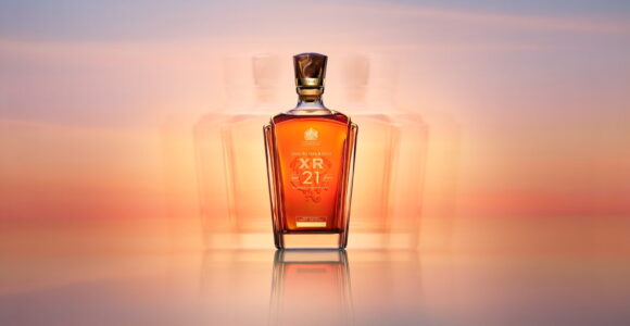 A richly colored bottle with a square base tapers upward to a narrow neck, capped by a dark stopper. The glass reflects golden hues, set against a softly blurred background of pink and orange gradients, reminiscent of a serene sunset. The bottle's elegant curves and warm tones suggest luxury and refinement, evoking a sense of sophistication.