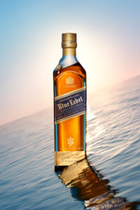 A tall, rectangular bottle with golden liquid inside stands on a shimmering, watery surface, catching the warm light of a setting sun. The blue and gold label contrasts elegantly with the amber contents. Reflecting on the gentle ripples, the bottle's cap is adorned with intricate details, enhancing its premium appearance against a serene, gradient sky.