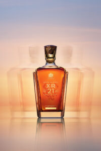 A sophisticated bottle of amber liquid stands elegantly against a serene, gradient background of soft blues and warm, peachy tones. The glass bottle features a distinct, ornate design with a reflective surface that mirrors the colors above. Flanking the central bottle, blurred reflections create a subtle, dynamic balance. The composition exudes luxury and refinement.