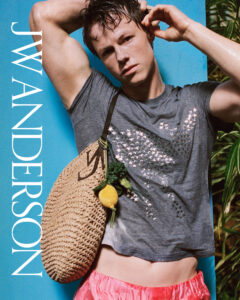 A young man leans against a bright blue surface, his damp hair and skin suggesting heat or recent activity. One arm lifts behind his head while the other adjusts his ear, creating a relaxed yet styled pose. He wears a fitted grey t-shirt with a subtle pattern, loose red shorts, and carries a woven bag adorned with a small decorative charm, evoking a fresh, summery mood.