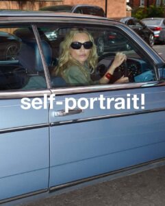 A person with long blonde hair, wearing sunglasses and a dark bracelet, sits behind the wheel of a classic blue car. The vehicle is parked on a street, with other cars visible in the background. The person, wearing a green top, looks out of the open window, with both hands on the steering wheel, conveying a relaxed yet attentive demeanor.