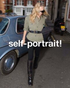 A person with long blonde hair and sunglasses walks confidently down a street beside a classic blue car. They wear a belted olive green jacket, black pants, and tall black boots. The background reveals an urban setting with brick buildings and a parked motorcycle. The scene captures a casual yet stylish moment in a city environment.